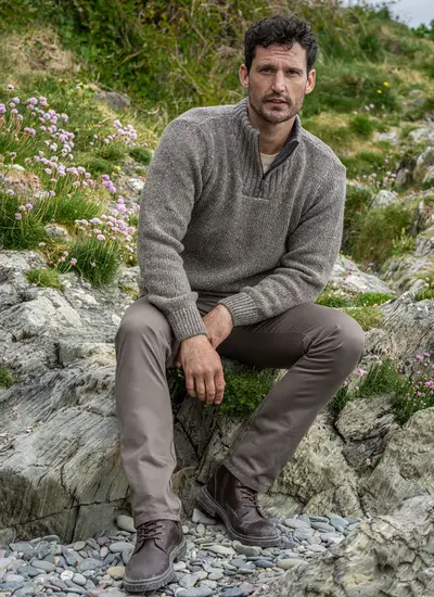 Full seated view of man in Glenstone Rocky Ground Sweater, styled with lace-up brown boots and coastal landscape backdrop.